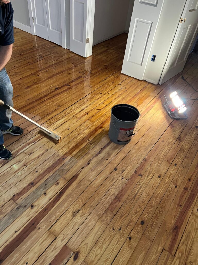 A technician from Precision Hardwood Flooring applying a clear, protective topcoat to a wide-plank pine floor in a Bergen County, NJ home.