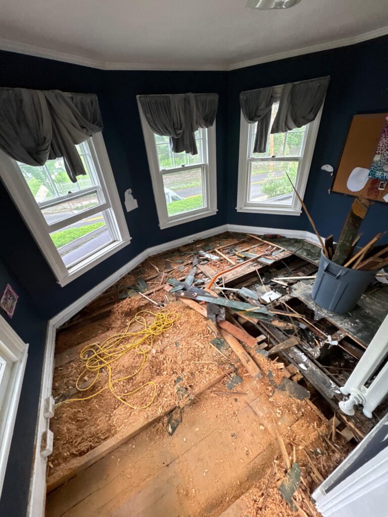 A residential room in Bergen County, NJ, undergoing complete floor demolition by Precision Hardwood Flooring, showing exposed subfloor joists and debris in a room with navy blue walls.