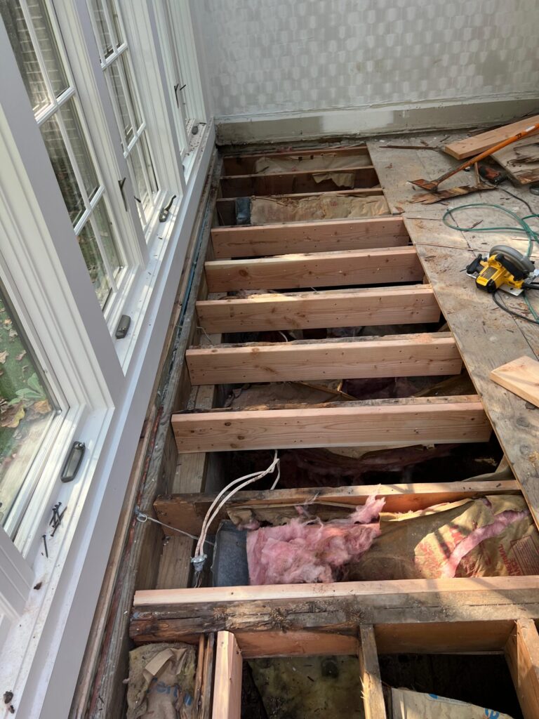 Residential floor renovation showing exposed joists, insulation, and subfloor removal along a windowed wall during hardwood flooring preparation.