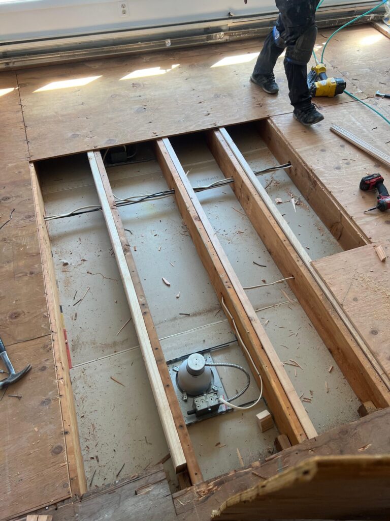 Exposed floor joists and open subfloor with electrical components visible during hardwood flooring installation preparation.